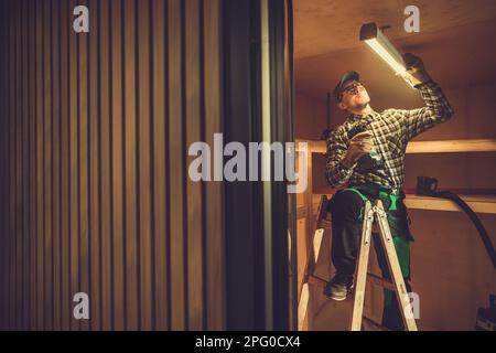 Caucasian Electrician in His 40s Installing LED Light Fixture in the Shed Ceiling. Small Wooden Building Illumination Stock Photo