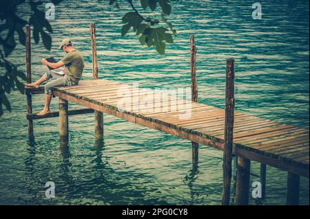 Computer Remote Work While on Vacation. Caucasian Man Using His Computer in Front of Scenic Lake. Seating on Wooden Pier Deck. Stock Photo