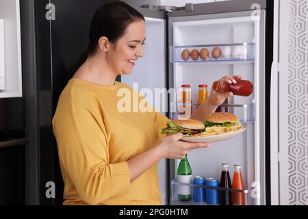 Happy overweight woman with ketchup and burgers near fridge in kitchen Stock Photo