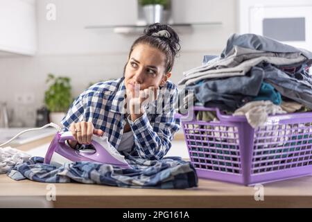 Woman is daydreaming while ironing next to an overfilled big basket full of clothes. Stock Photo