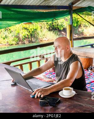 Working remotely on his computer at Si Phan Don,a man traveling the world,sits on a balcony at a rural guesthouse,overlooking the Mekong river,with a Stock Photo