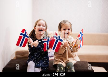 Two sisters are sitting on a couch at home with norwegian flags on hands. Norway children girls with flag . Stock Photo