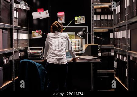 Woman private detective studying crime case board hanging on wall. African american police investigator searching solution while analyzing clues and murder scene photograph at night Stock Photo