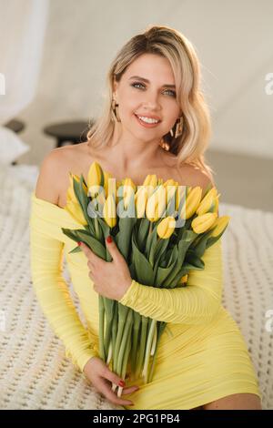a happy woman in a yellow dress embraces a bouquet of yellow spring tulips in the interior. Stock Photo