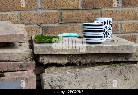 Mugs, screws, artificial grass and scissors on a pile of dirty paving slabs in front of a brick wall. Stock Photo