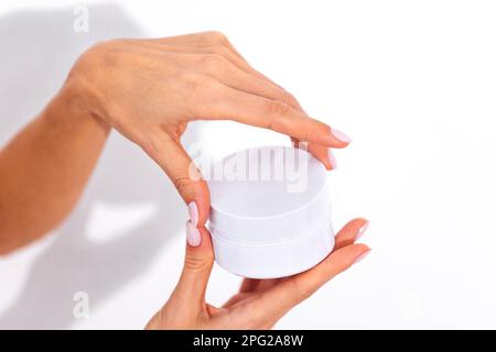 Extreme close up of beautiful graceful female hands with white manicure holding mock up of round box for beauty product. White background. Isolated. F Stock Photo