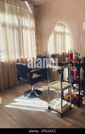 Empty work place for hairdresser in beauty salon. Comfortable black swivel chair for the client and large shaped mirror. Open shelves on wheels with c Stock Photo