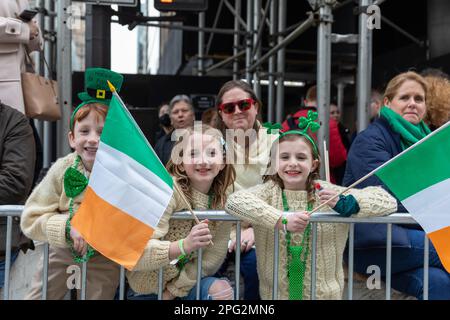 Three joyful young girls wearing shamrock-patterned glasses and waving Irish flags stand in front of a metallic fence Stock Photo