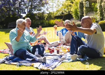 Happy group of diverse senior friends having picnic and talking in garden on sunny day Stock Photo