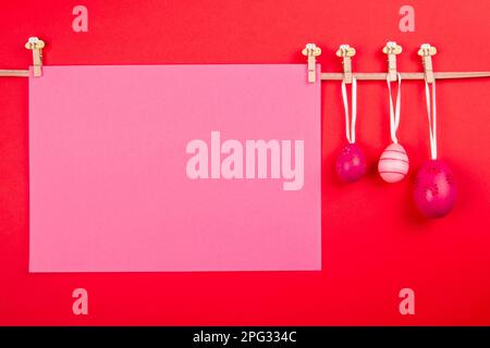 Blank rose colored sheet of paper and three pink Easter eggs hang at rope on red background. Easter card template featuring blank area for text Stock Photo