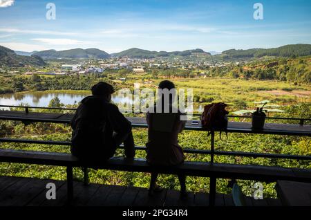 A young Vietnamese couple sit at the Me Linh Coffee Garden and look out across coffee trees, Dap Cam Ly Lake and a valley in the central highlands nea Stock Photo