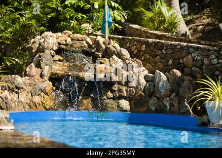 An artificial, small waterfall that flows into a basin on stones in Coron in the Philippines surrounded by tropical plants. Stock Photo