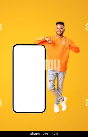 Handsome young 20s middle-eastern man standing in full length near huge smartphone with empty screen and showing thumb up, mockup, handsome indian guy recommending mobile app, collage Stock Photo