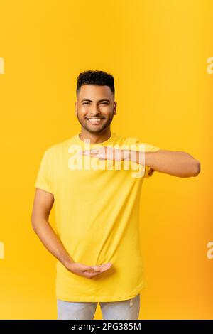 Positive Indian man looking at camera, holding two hands in front of him showing height size of something, wearing casual t-shirt, smiling joyfully, presenting product standing isolated on yellow Stock Photo