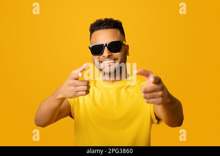 Hey you. Excited positive young arab man in sunglasses pointing fingers at the camera, choosing you, handsome indian guy looking at the camera and laughing, isolated on yellow background Stock Photo