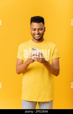 Photo of attractive young 20s indian man in casual t-shirt using smartphone isolated over yellow background, touches phone screen, necessary tool for productivity, finds inspiration online Stock Photo