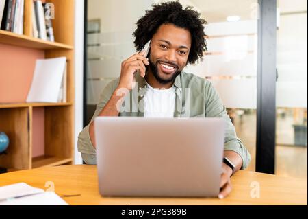 Friendly and joyful african-american guy making phone call and working with laptop indoors, male office employee talking on the phone during break, salesman staying in touch with clients Stock Photo