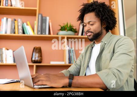 Friendly and positive african-american male employee or student using laptop, businessman looking at the screen, typing, responding to emails sitting in contemporary office Stock Photo