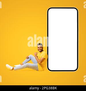 Excited handsome man points finger at huge smartphone with empty blank screen sitting near on the floor isolated on yellow, happy indian guy presenting mobile app or best deal, copy space, template Stock Photo