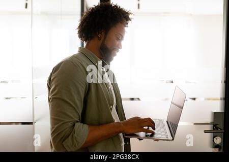 Concentrated man typing and working on a laptop remotely, standing in modern coworking space, focused and serious african-american freelancer, male office employee checking emails, side view Stock Photo