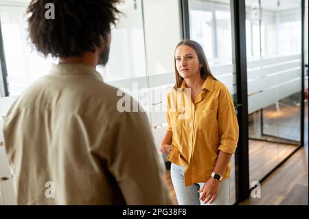 Two coworkers chatting during coffee break standing in glass hall of office building. Curly african-american male employee explain something for female colleague, sharing his ideas, woman listening Stock Photo