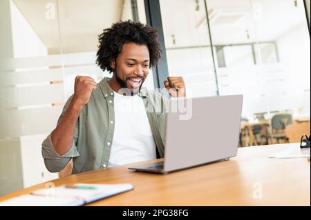 Happy african-american curly male freelancer excited about job offer, raising hands and laughing, man celebrating successful deal or online win, screaming yes, lucky guy received good news by email Stock Photo