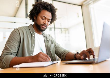 Serious concentrated young african-american businessman, ceo, manager working on laptop, doing online market research, working on project, creating plan or checking financial statement, taking notes Stock Photo