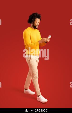 Waist up view of happy handsome blogger young adult man standing, using smartphone and texting message with smile. Indoor, isolated, studio shot, red background, copy space Stock Photo