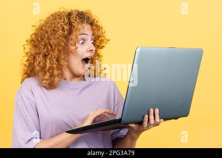 Studio image with yellow background of a beauty surprised woman with curly hair using a laptop while standing Stock Photo