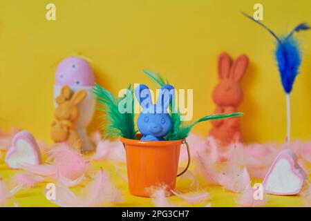 a blue easter bunny in an orange bucket surrounded by pink feathers and a pink easter egg and some other easter bunnies of different colors on a yello Stock Photo
