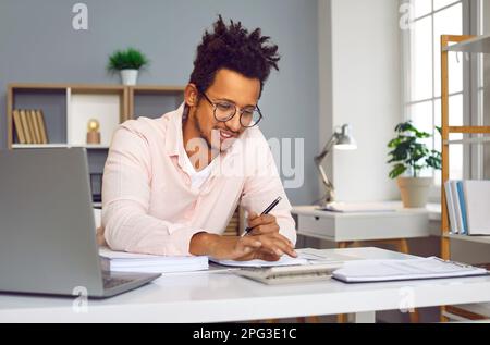 Young business man working on laptop at office, smiling and making calculations Stock Photo