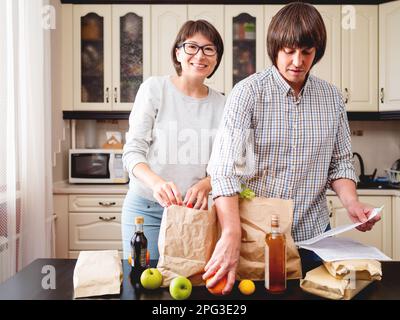 Young couple is sorting out purchases in the kitchen. Products in bags made of craft paper. Food delivery. Stock Photo