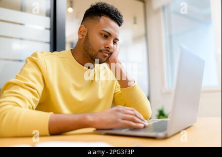 Bored Indian male student feels lack of motivation, office employee feeling weary because lacks interest in current activity, sitting in front of laptop, resting chin with hand Stock Photo