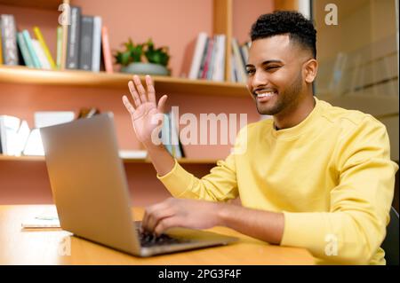 Smiling Indian man, office employee or student in casual clothes sitting at the desk, studying or working online, having a virtual meeting, saying hello, waving at the laptop screen Stock Photo