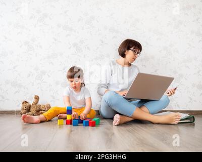 Mom and son at home. Mother works remotely with laptop and smartphone, son plays with toy blocks. Stock Photo