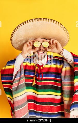 Fresh. Young funny man in colorful festive clothes, poncho and sombrero posing with lime near eyes against yellow studio background Stock Photo