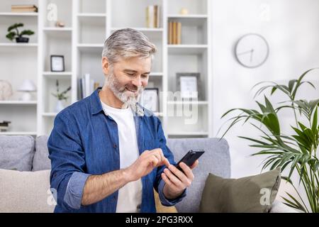 Mature adult man close-up at home smiling and using phone in living room sitting on sofa, person typing message and browsing web page. Stock Photo