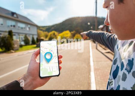 A man's hand holds a smartphone with an online map, and a teenager points the way with his hand. In the background is a city street with an empty road Stock Photo