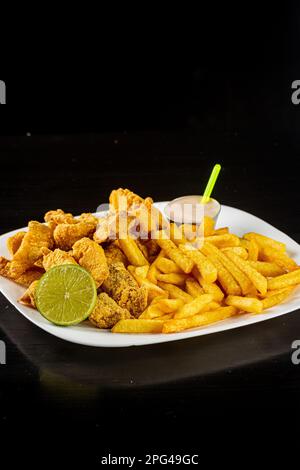A white plate containing freshly-fried potato fries and chicken nuggets, garnished with a wedge of lime and a sauce Stock Photo