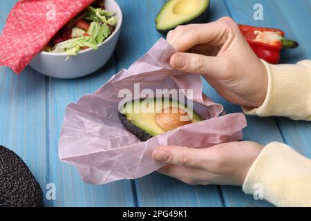 Woman packing half of fresh avocado into beeswax food wrap at light blue wooden table, closeup Stock Photo