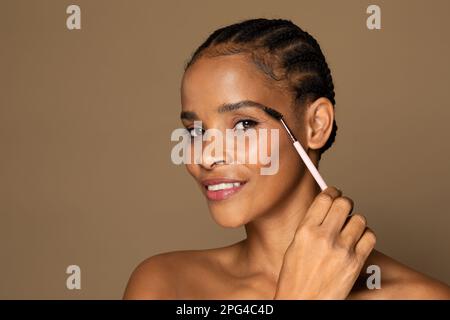 Natural makeup. Attractive middle aged black woman brushing eyebrow holding brush, brown background, free space Stock Photo