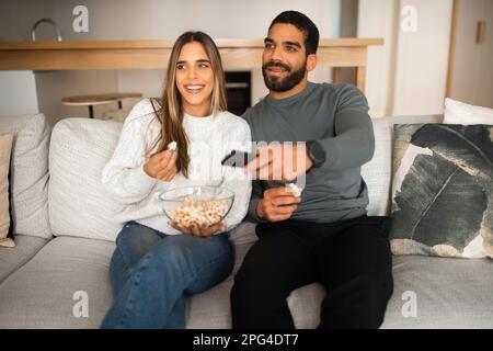 Laughing multiracial young couple sit on sofa, enjoy free time together, watch tv with remote control, eating popcorn Stock Photo