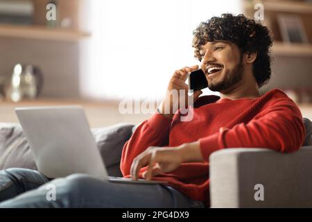 Happy Indian Man Talking On Cellphone And Using Laptop At Home Stock Photo