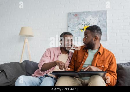 Smiling african american man pointing at photo album near shocked son at home,stock image Stock Photo