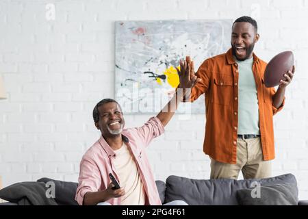 Positive african american father and son giving high five while watching rugby match at home,stock image Stock Photo