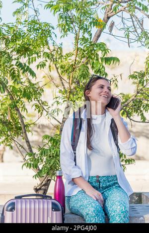 Young woman smiling while talking on the phone sitting outdoors with her suitcase. Stock Photo