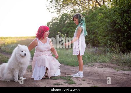 Girl with green hair gives wild flowers to mother with pink. Traveling with Samoyed dog, pets to nature. The tenderness of motherhood, one parent. Div Stock Photo