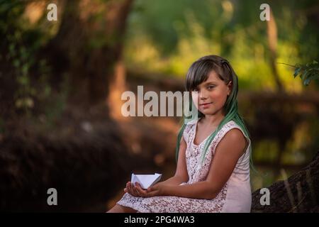 Portrait of girl with green hair in vintage dress, sits on the river bank, holding white origami paper boat in hands. Child cheerfully launches the sh Stock Photo