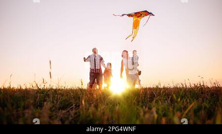 Panoramic diversity view of family running at sunset with kite in the sky. View from below through the ears. Silhouette. The father holds toy in hands Stock Photo