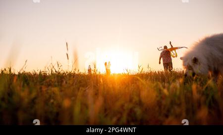 Defocused panoramic diversity family with Samoyed running at sunset with kite in sky. View from below through the ears. Silhouette. Father holds toy i Stock Photo